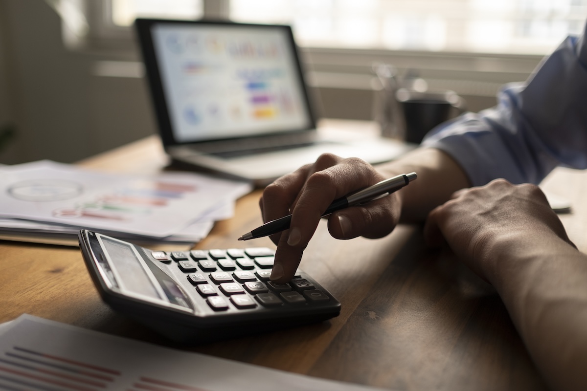 A man’s hand using a calculator, placed on a wooden desk with financial papers and a laptop in the background. Calculating required minimum distributions.